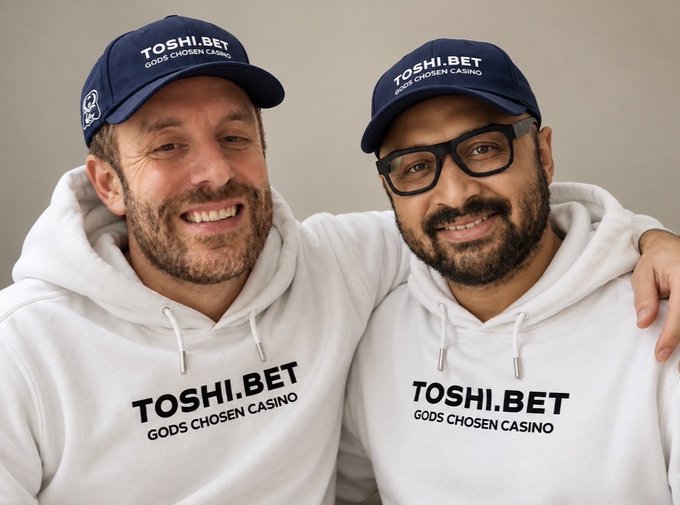 Two smiling men wearing matching navy 'TOSHI.BET' caps and white 'TOSHI.BET GODS CHOSEN CASINO' hoodies standing side-by-side.