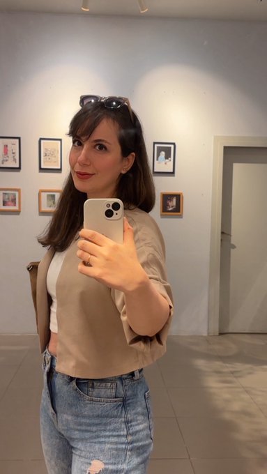 A woman takes a mirror selfie in an art gallery, wearing a tan blouse, white top, and jeans, with sunglasses perched on her head.