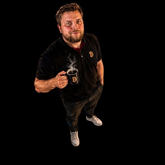 A man with a beard wearing a black polo shirt with a Bitcoin logo and dark pants, holding a black mug with a Bitcoin symbol and steam rising, against a black background.