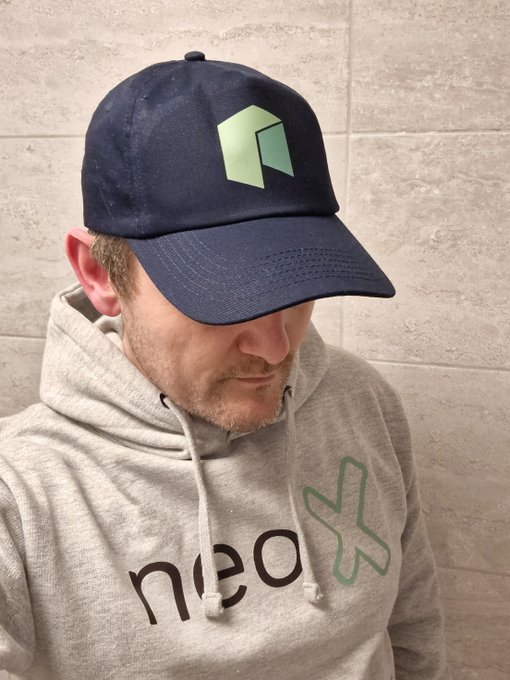 A man wearing a navy cap with a green logo and a grey 'neo' hoodie looks downward.