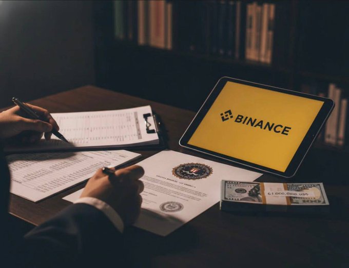 A person's hands writing on documents at a wooden desk, with a tablet displaying the "BINANCE" logo in yellow, a stack of US hundred-dollar bills, and financial paperwork with a logo seal.