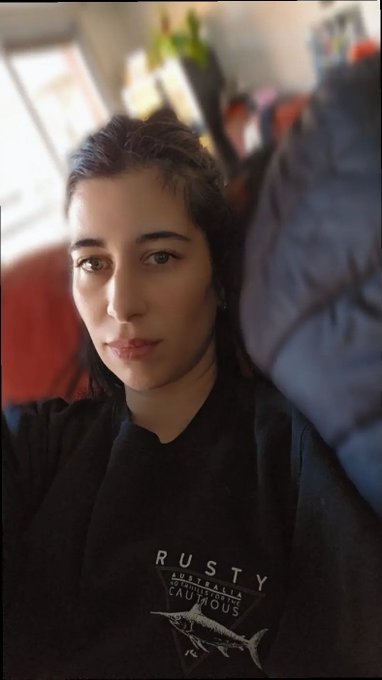 A selfie of a young woman with dark hair and freckles wearing a black "Rusty Australia" sweatshirt, sitting on a couch.
