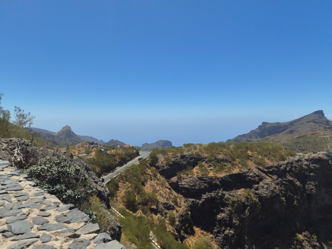 A scenic mountain landscape with a winding road descending through rocky, brush-covered terrain toward the ocean under a clear blue sky.