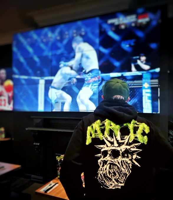 A person wearing a green cap and a black hoodie with neon green 'AYEPE' text and a spiked skull graphic watches a UFC fight on a large TV screen in a dimly lit room.