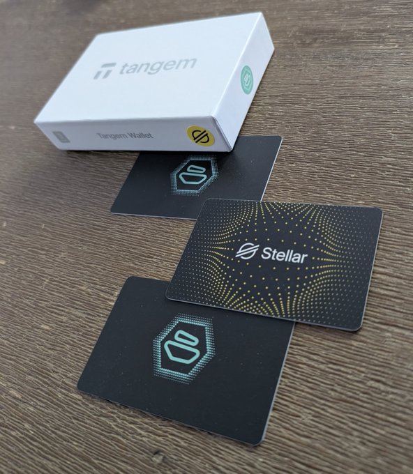 A Tangem Wallet box sits on a wooden surface next to two cards, one displaying a logo and another featuring the Stellar cryptocurrency branding with a gold dot pattern.