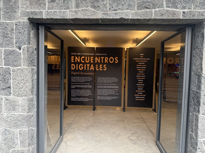 An entrance to an exhibition featuring black informational panels with orange text reading 'ENCUENTROS DIGITALES / Digital Encounters', displaying the names of participating artists.