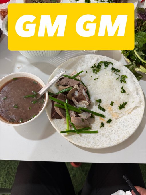 Overhead photo of a Vietnamese meal with a bowl of dark broth and a plate of rice with meat and herbs, overlaid with a yellow banner reading 'GM GM'.