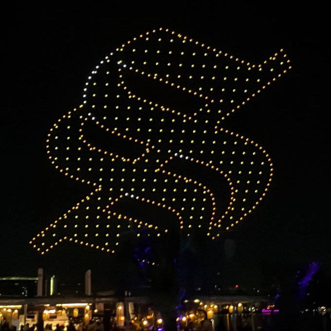 A massive drone light show forms a glowing orange/orange-tinted Solana logo in the night sky above a waterfront, with buildings and crowds visible below. The display likely commemorates the speaker's favorite Solana moment—Breakpoint 2025 in Abu Dhabi—capturing the scale and spectacle of the conference.