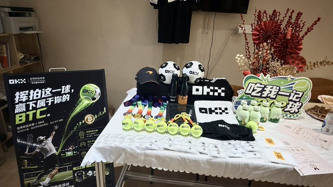 A promotional display table for OKX features branded merchandise including tennis balls, soccer balls, baseball caps, and tote bags, alongside a poster reading "挥拍这一球,赢下属于你的 BTC" (Swing this shot, win your BTC) and "吃我一记球" (Take my shot), tying into the caption's mention of learning tennis, golf, soccer, and F1 to engage clients with sports-themed crypto incentives.