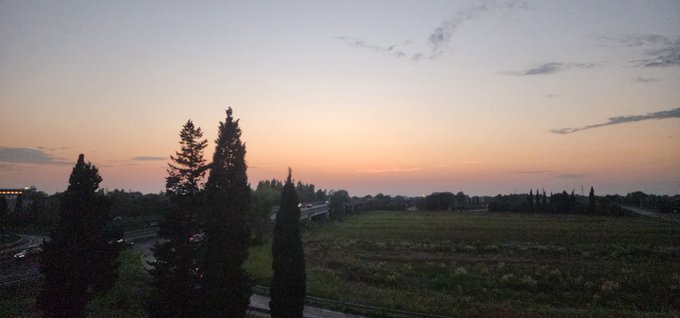 A panoramic sunset view over a countryside landscape with tall silhouetted trees in the foreground, a field, and distant buildings under a gradient sky of orange and blue.