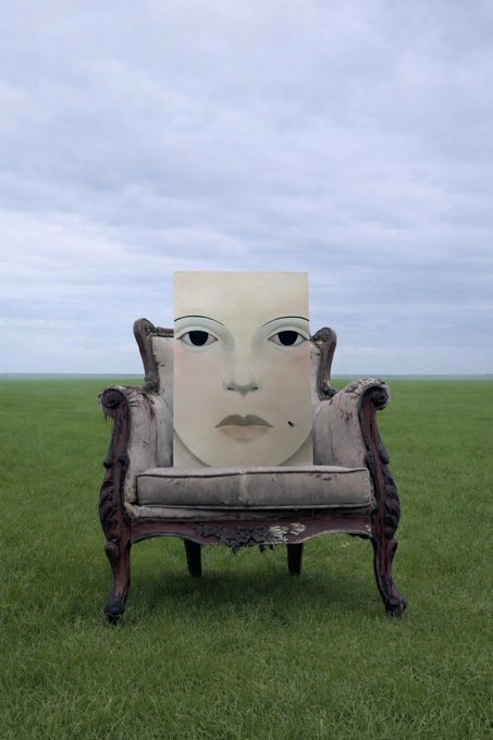 An antique ornate armchair sits in a grassy field under a cloudy sky, with a large canvas painting of a face propped on its seat.