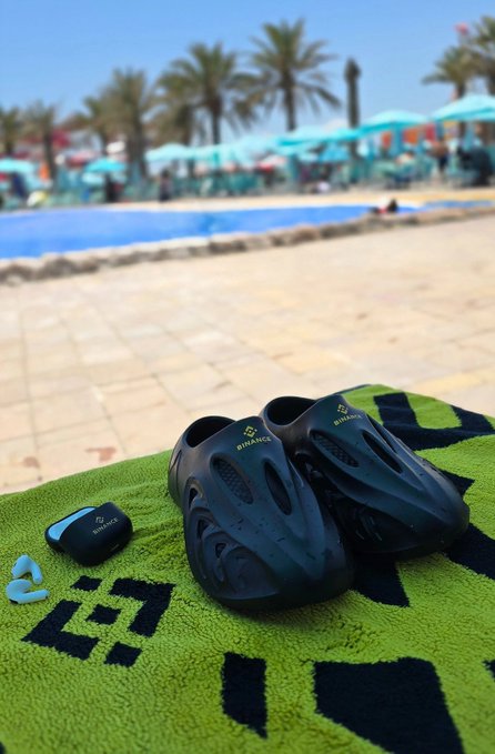 A pair of black Birkenx slides and matching Birkenx Bluetooth earbuds rest on a green towel by the pool, capturing the perfect "summer setup is elite fr" vibe with palm trees and lounge chairs in the background.