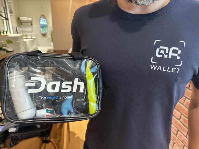 A person wearing a navy blue 'RR Wallet' t-shirt holding a clear toiletry bag with 'Dash - The world is home' branding, containing various travel items, photographed in what appears to be a bathroom setting.