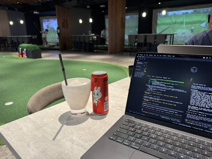 A workspace setup featuring a laptop displaying code on the screen, accompanied by a glass of a milky beverage with a straw and a red canned drink, all placed on a table overlooking an indoor golf simulator facility.