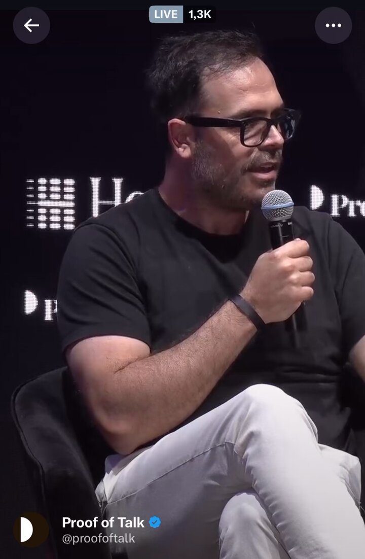 In this image, a man wearing glasses and a black shirt is sitting down while holding a microphone. The background of the image features a sign that says "Hilton Paris" with a logo for "proofoftalk". The caption below the image reads "@kostascrypto @proofoftalk Paris", which suggests that the photo was taken at a live event or discussion, possibly related to the agentic economy and Sui's object-centric scalable model.