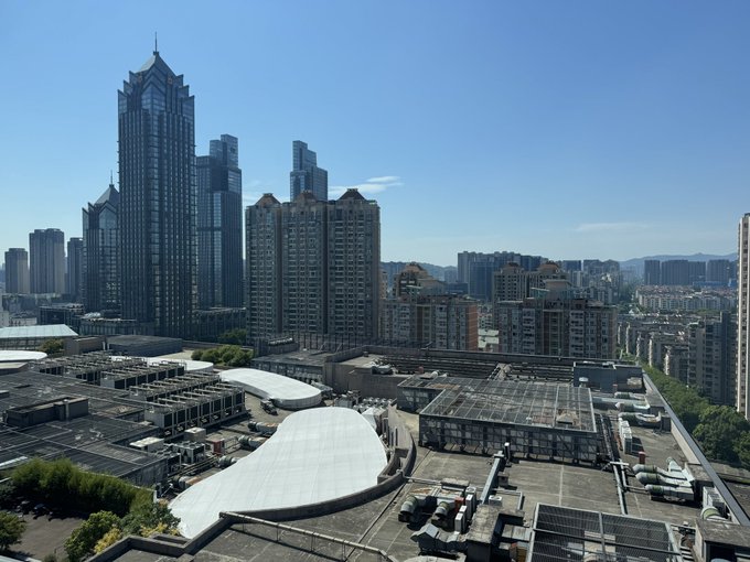Daytime cityscape showing modern high-rise buildings and skyscrapers in the distance with industrial rooftops in the foreground under a clear blue sky.