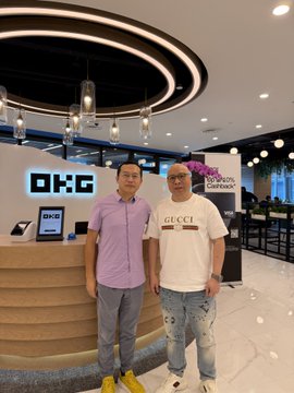 Two men pose together in front of an "OKX" branded reception desk, with the man on the right wearing a Gucci t-shirt. The image relates to the post about meeting @star_okx, showing the person looking healthy and youthful, while the caption references an older photo suggesting improvement over 12 years.