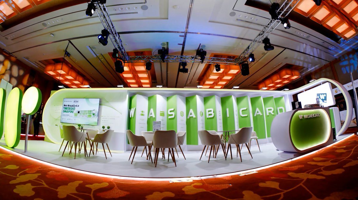A wide-angle view of a brightly lit WASABI CARD exhibition booth or lounge area, featuring green vertical panels with the company's name, modern white furniture, a presentation screen, and an elaborate ceiling rig with stage lights.