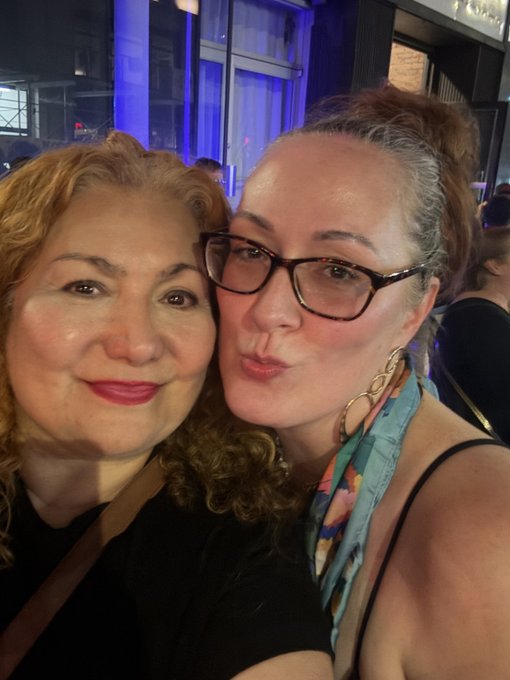 A selfie of two women posing closely together, one with curly hair and one wearing glasses and a colorful scarf.