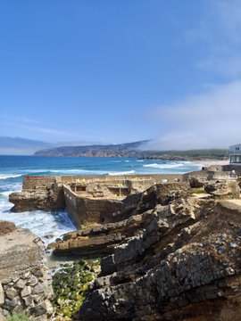 In the image, there's a view overlooking a rocky beach area with large rocks protruding from the water. The background appears to be an ocean or a large body of water. On this picturesque landscape, there are several people enjoying their time near the shoreline and along the coast as well. The presence of these individuals suggests that it might be a popular location for leisure activities such as sunbathing, swimming, or walking along the beach.