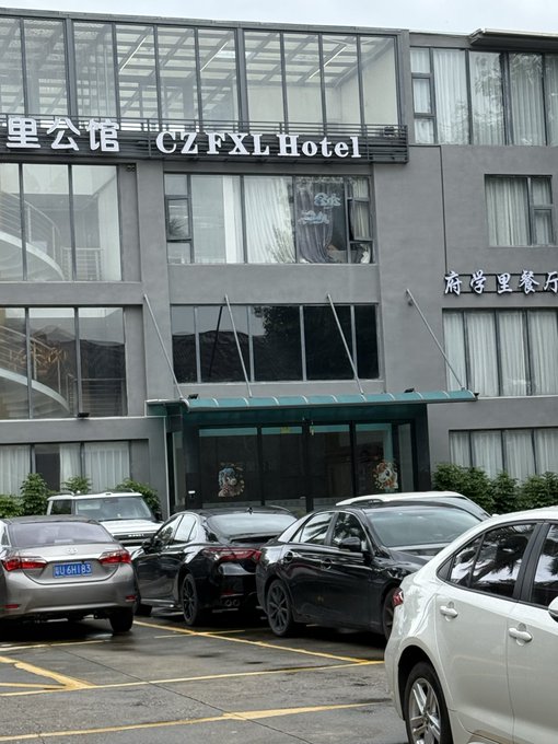 A photo of the "CZ FXL Hotel" entrance, with cars parked in front of the grey building. The caption humorously plays on the hotel's name referencing "CZ," matching the post's joke about CZ (Changpeng Zhao, Binance's founder) apparently opening his own hotel.