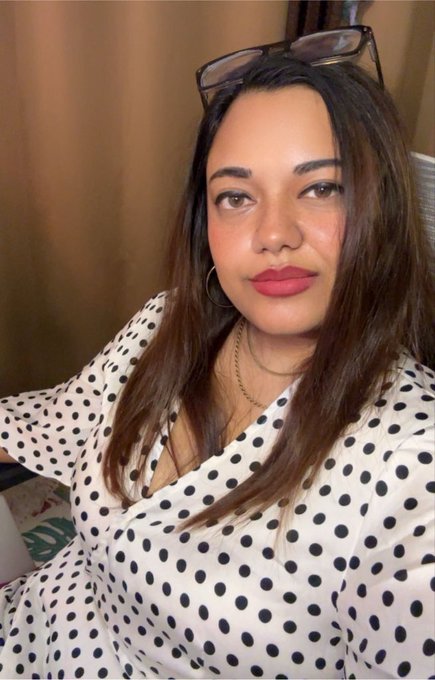 A selfie portrait of a woman with long brown hair, glasses perched on her head, hoop earrings, and pink lipstick, wearing a white blouse with black polka dots.