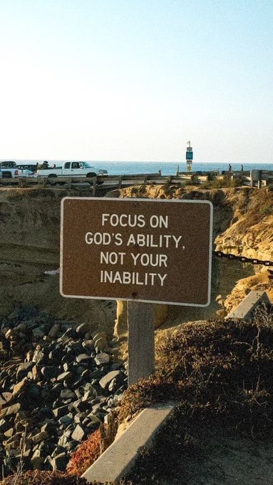 A photograph of a brown sign on a coastal cliff reading 'FOCUS ON GOD'S ABILITY, NOT YOUR INABILITY', with a lighthouse and ocean visible in the background.