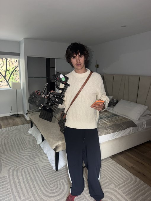 A young man with dark wavy hair stands in a modern bedroom, holding a professional camera rig with a microphone while wearing a cream sweater and black track pants. The caption humorously suggests this is someone named "youngjazzeth" who has unexpectedly appeared in their house, jokingly asking "a1lon9" for help with the unexpected visitor.