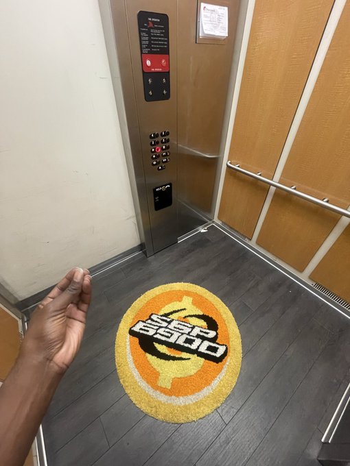 Inside an elevator, a hand reaches toward a round orange doormat featuring the 'Super Scandal' logo.
