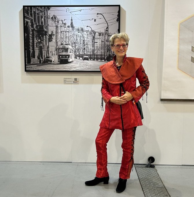 The image features an older woman wearing red clothing, standing next to a painting of a cityscape with a bus passing by. The woman appears to be posing for a photo at the gallery. The painting depicts a scene from Prague, which is the location where the gallery is located. The artwork is titled "Timeshifting Prague" and it is part of a series that explores the concept of time and its impact on our lives.