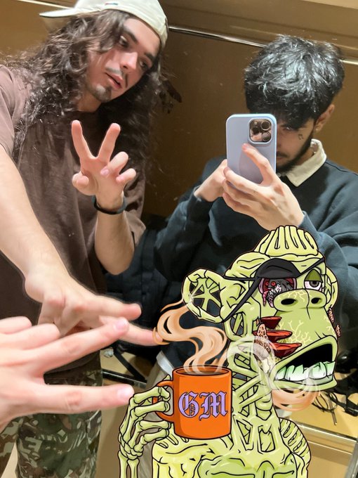 Two young men taking a mirror selfie, one making a hand gesture, with a cartoonish green skeletal character holding a steaming mug labeled 'GM' overlaid at the bottom.