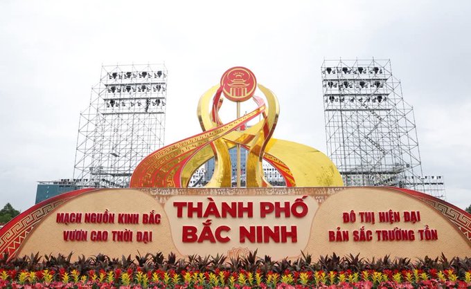 A large welcome monument or sign reading 'THÀNH PHỐ BẮC NINH' (Bac Ninh City) flanked by slogans in Vietnamese, decorated with flowers and featuring a golden emblem, with metal scaffolding behind it.