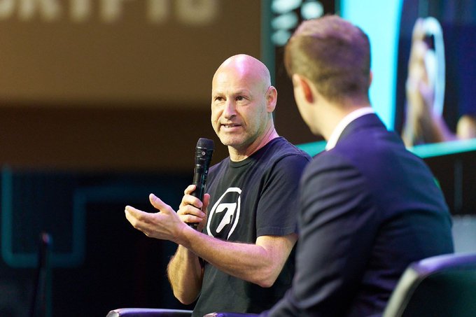 The image shows a man wearing a black shirt holding a microphone, talking to another person who is also wearing a suit. The background of the scene includes a TV screen displaying an advertisement for Bitcoin. The text "FUD Check: The recent Ethereum Foundation cuts & departures are NOT a crisis" and "Consensys." suggests that there might be some discussion or disagreement about the current state of the Ethereum Foundation, which has been facing criticism for its focus on core protocol tech rather than commercialization.