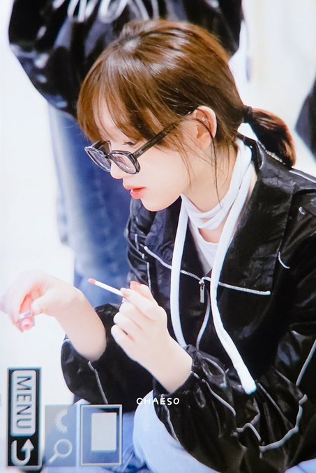 A photograph of a young woman with glasses and a black jacket, looking down at a small object in her hands, with 'MENU' interface text visible.