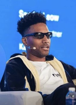 A man with curly hair and sunglasses speaks into a headset microphone at what appears to be a conference, with partial text "SN" and "LON" visible in the background. The image accompanies the post about crypto trader Ansem declaring that the current setup is the best for a crypto bull market in a very long time and urging people to lock in now for potential life-changing gains in the next two years.