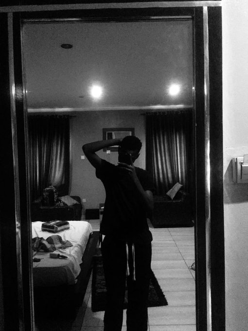 Black and white mirror selfie of a person in a dimly lit hotel room, with a bed and dark curtains visible in the background.