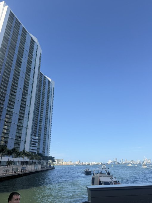 A view from a boat near tall waterfront high-rises on a clear blue day, with several boats visible on the water in the distance. The image captures the casual, on-the-water setting referenced in the post about being on a boat in Miami.