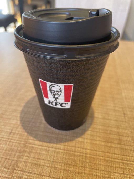 A close-up photograph of a brown paper hot beverage cup bearing the KFC logo, sealed with a black plastic lid, sitting on a wooden table.