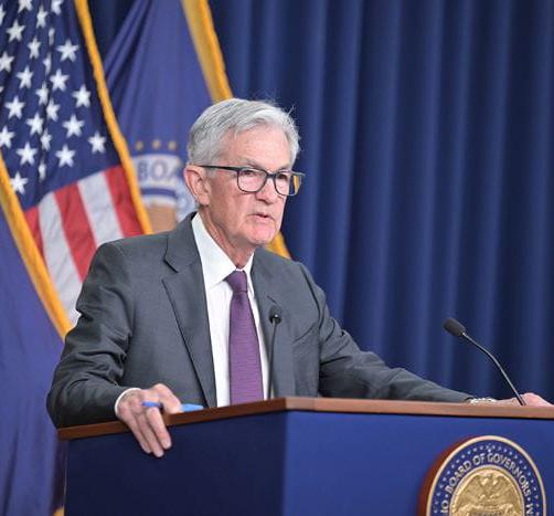 Federal Reserve Chair Jerome Powell stands at a podium during a press conference, with an American flag and the Board of Governors seal visible.