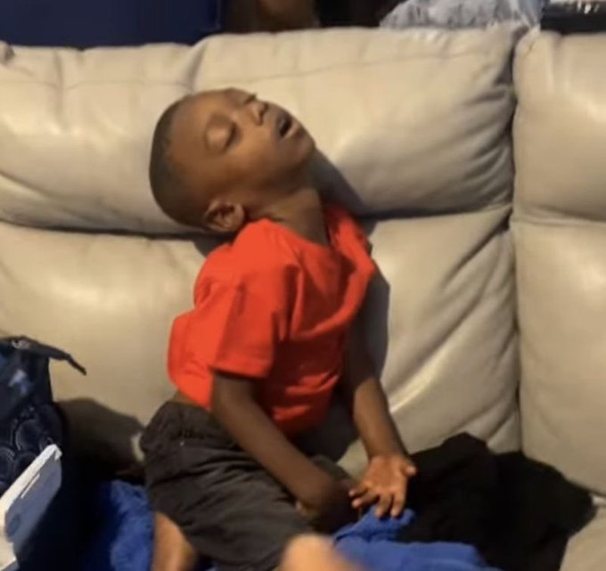 A young boy in a red shirt sits on a white couch, head tilted back and mouth open as if sleeping or exhausted.