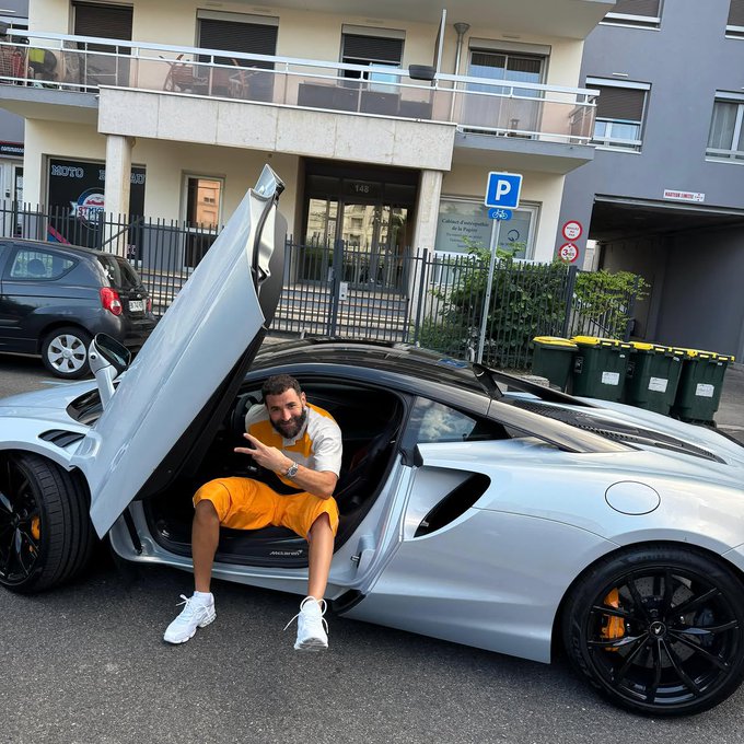 A man in an orange outfit sits in the open butterfly door of a silver McLaren sports car parked on a street, making a peace sign, with a building and 'P' parking sign visible in the background.