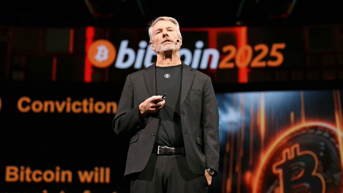 The image features an older man wearing a suit, standing on stage at a conference or event. He appears to be giving a speech or presentation about Bitcoin. The background of the image shows a large screen displaying the Bitcoin logo with the text "Bitcoin takes a hit as major holder breaks with 'never sell' mantra tijd.be/markten-live/n …" This suggests that there might have been an incident involving Bitcoin, possibly related to its value or market sentiment.