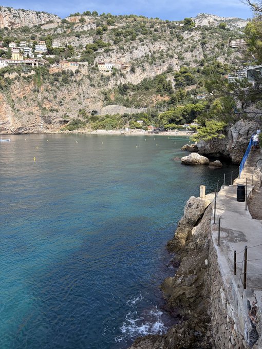 Crystal clear Mediterranean waters lap against a rocky cove on the Côte d'Azur, with hillside villas overlooking a small beach and a stone walkway hugging the cliff edge.