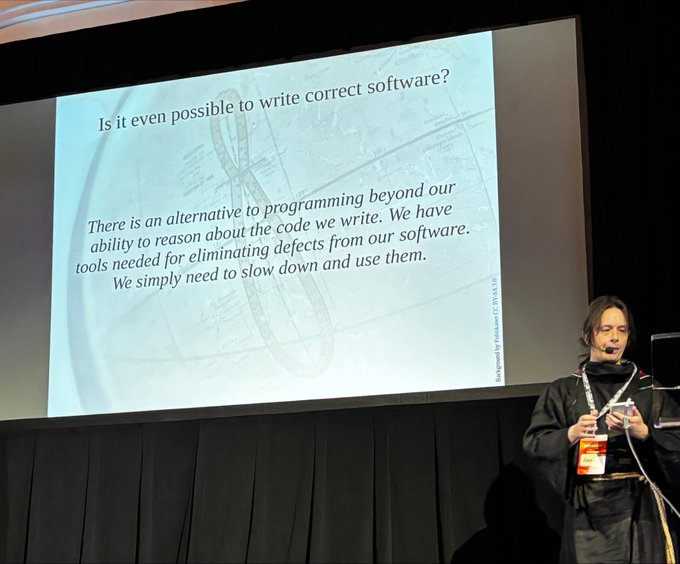 A presenter on stage at a conference standing next to a slide titled 'Is it even possible to write correct software?' with text discussing tools for eliminating software defects.
