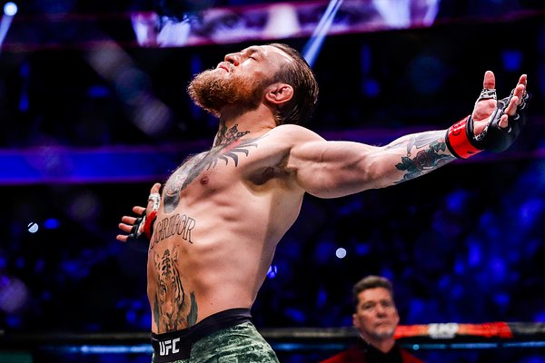 A shirtless, tattooed UFC fighter stands in the octagon with arms raised triumphantly toward the ceiling, his head tilted back in celebration.