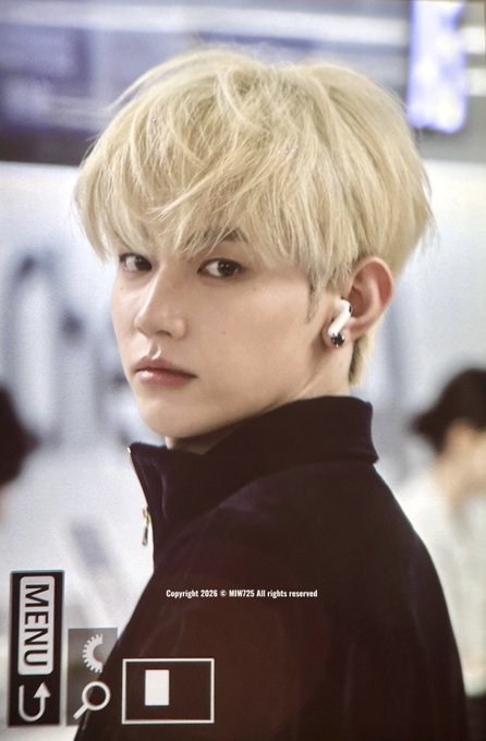 A person with blonde hair wearing a dark jacket and white earbuds, looking over their shoulder at the camera in what appears to be an airport setting. The image features a copyright watermark reading "© MIMI25 All rights reserved" along with camera UI elements like "MENU" and a record button, consistent with a candid fancam taken during an airport arrival as referenced in the post about Zhang Hao's GMP airport appearance.