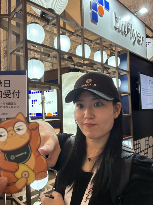 A young woman wearing a black "MANTLE" branded cap poses at the booth of Mantle partner Bitflyer during WebX2026, holding an orange Maneki-neko (lucky cat) mascot also branded with the Mantle logo. The event's networking scene is captured with Bitflyer's illuminated lantern-decorated signage and Japanese text regarding audio reception in the background.