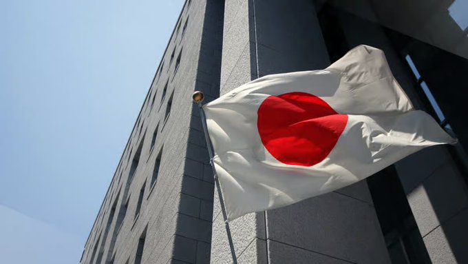 A Japanese flag (white with a red circle) waving on a flagpole in front of a modern building against a clear blue sky.