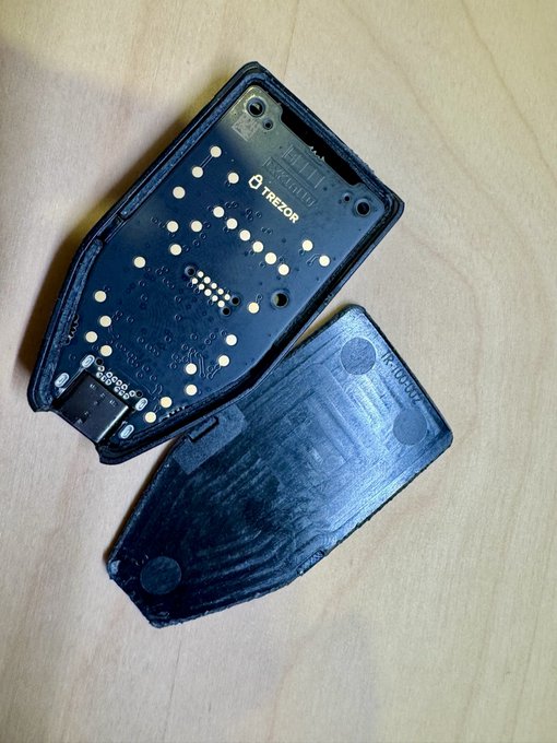 A disassembled Trezor hardware wallet showing its PCB (labeled "TREZOR") and detached back cover, illustrating the post's point about Trezor's tamper-evident one-piece shell design that differs from snap-fit or glued alternatives—once opened, the separated pieces are clearly visible on the wooden surface.