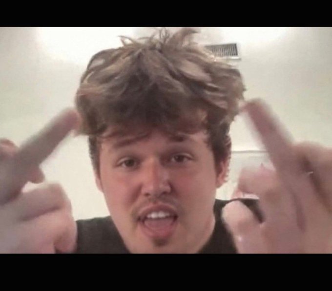 A young man with tousled hair and a goatee points both index fingers toward his head while speaking directly to the camera in an indoor setting.